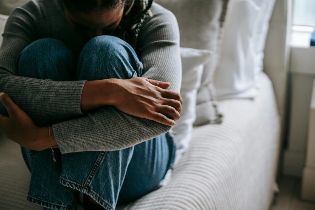A woman sits on a bed, hugging her knees in a contemplative pose, indoors.